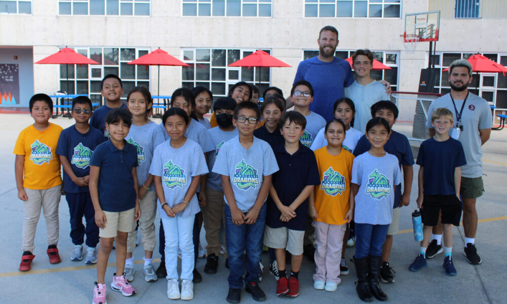 Chris Sale, a professional baseball pitcher, poses outdoors with a group of enthusiastic students from RCMA’s Immokalee Community Academy (ICA). The students, proudly wearing ICA-themed t-shirts and casual clothing, stand together smiling, with staff members and Sale in the back row. The vibrant scene takes place in front of the school building, with red patio umbrellas and a basketball hoop in the background, highlighting the engaging and welcoming environment of RCMA’s Immokalee Community Academy.