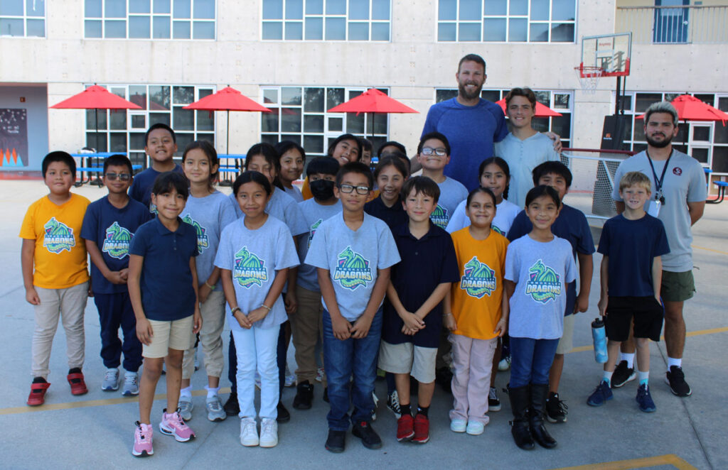 Chris Sale, a professional baseball pitcher, poses outdoors with a group of enthusiastic students from RCMA’s Immokalee Community Academy (ICA). The students, proudly wearing ICA-themed t-shirts and casual clothing, stand together smiling, with staff members and Sale in the back row. The vibrant scene takes place in front of the school building, with red patio umbrellas and a basketball hoop in the background, highlighting the engaging and welcoming environment of RCMA’s Immokalee Community Academy.