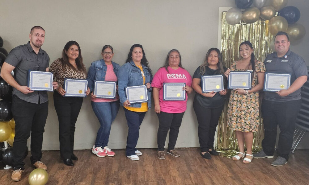 Group of RCMA Family Support Workers standing together indoors, holding certificates of completion for the Family Development Credential Program. The group is smiling, with a backdrop featuring gold and black balloons and a golden tinsel curtain.