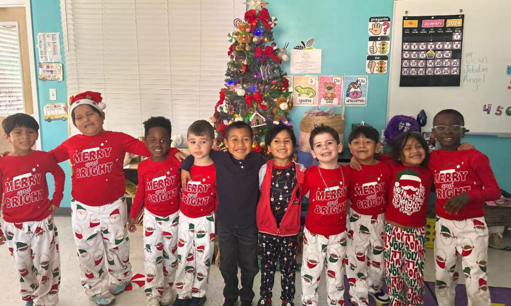 A joyful group of children from RCMA stands together in festive holiday attire, including matching red shirts that read 'Merry and Bright' and Santa-themed pajama pants. They smile brightly in front of a beautifully decorated Christmas tree adorned with ornaments, lights, and a shining star on top. The classroom background features cheerful wall decorations, educational posters, and a holiday calendar, creating a warm and festive atmosphere.